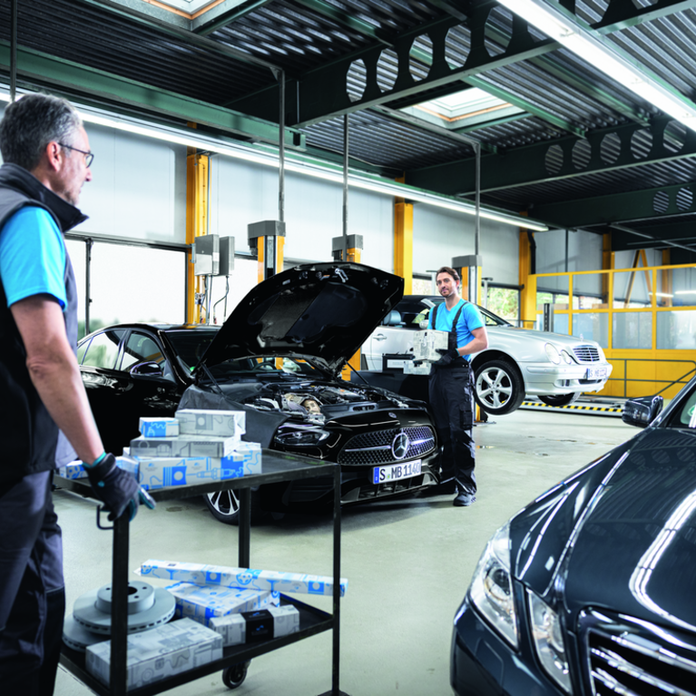 Mitarbeiter bei Ihrer Arbeit in der Mercedes-Benz Werkstatt. Einer der Mitarbeiter schiebt einen Wagen mit Ersatzteilen von Mercedes-Benz in die Werkstatt rein.